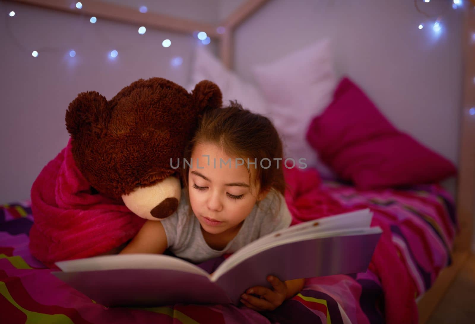 Its a story to spark the sweetest of dreams. a little girl reading a book in bed with her teddybear. by YuriArcurs