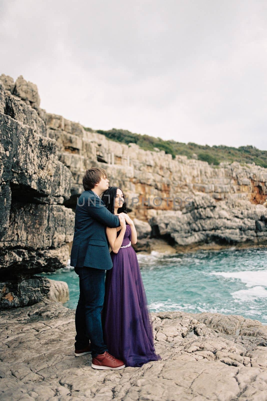 Man hugs woman from behind by the shoulders on the rocks by the sea. High quality photo