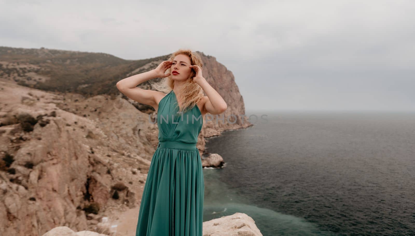 Side view a Young beautiful sensual woman in a mint long dress posing on a volcanic rock high above the sea during sunset. Girl on the nature on overcast sky background. Fashion photo