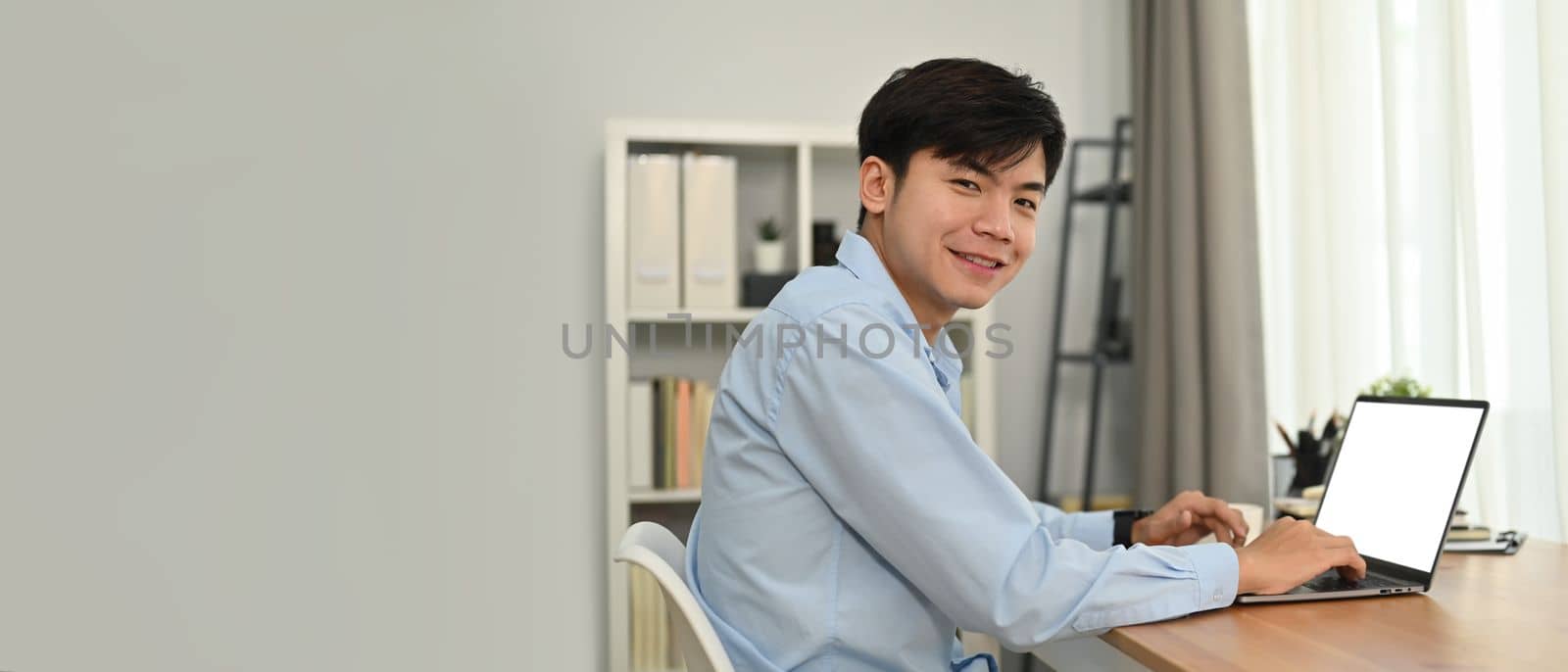 Cheerful asian man freelancer using laptop computer in modern home office. Horizontal banner, copy space for text.