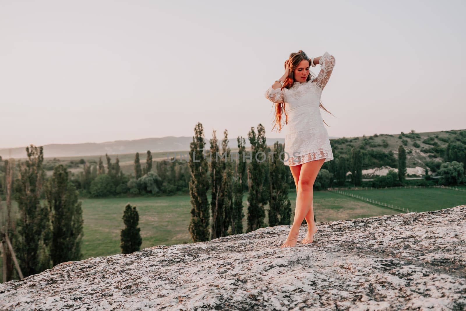 Happy woman in white boho dress on sunset in mountains. Romantic woman with long hair standing with her back on the sunset in nature in summer with open hands. Silhouette. Nature. Sunset. by panophotograph