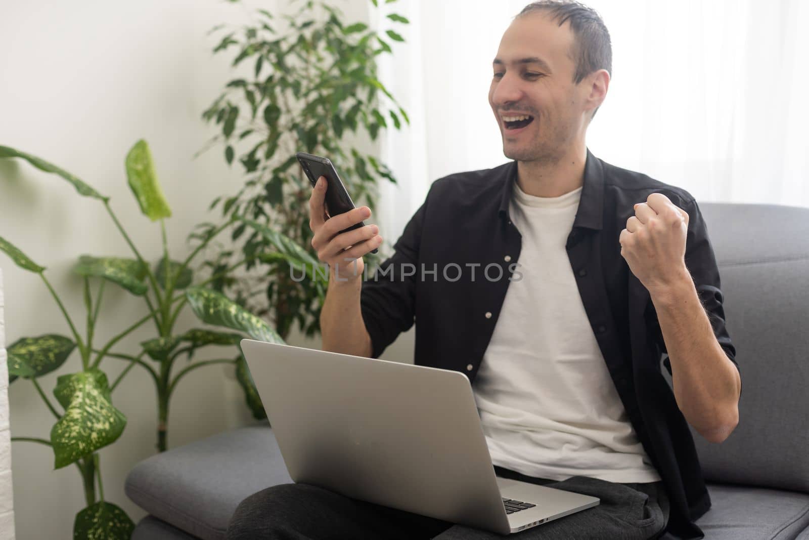 Smiling young man typing on mobile phone while sitting on a sofa at home with laptop computer.
