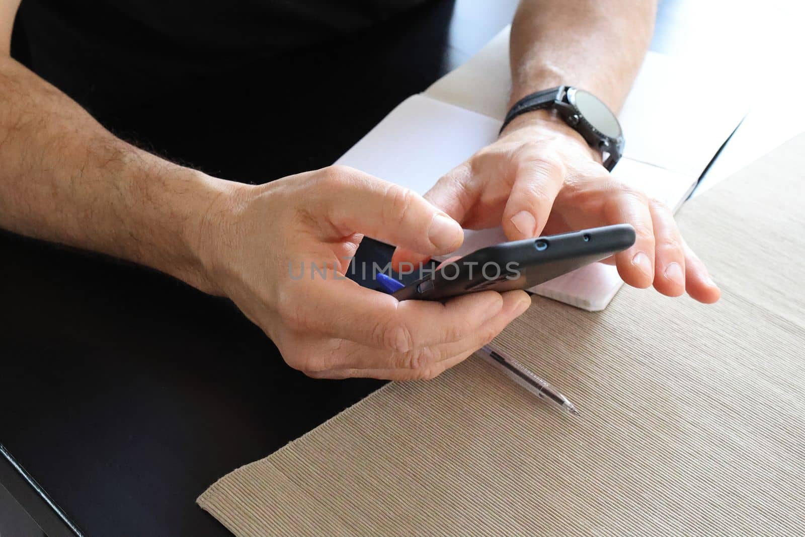 Close-up of the hands of a man with a smart watch on his hand, who is holding a black phone, looking for something. Black table with notepad and pen. Remote work concept by Proxima13