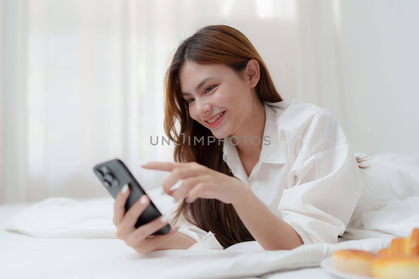 Beautiful asian woman on a white bed with coffee and using mobile phone at home. lifestyle concept by itchaznong