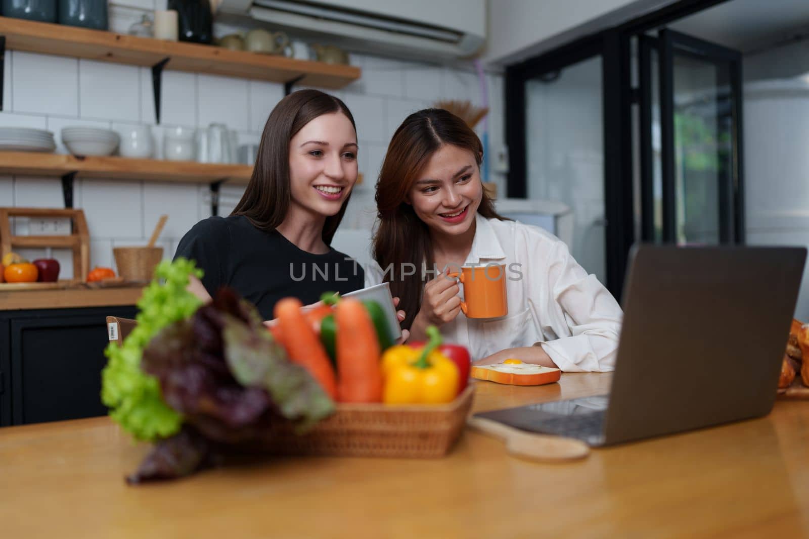 Beautiful woman and friend talking and using laptop at kitchen room. Girl friends couple living together at home. by itchaznong