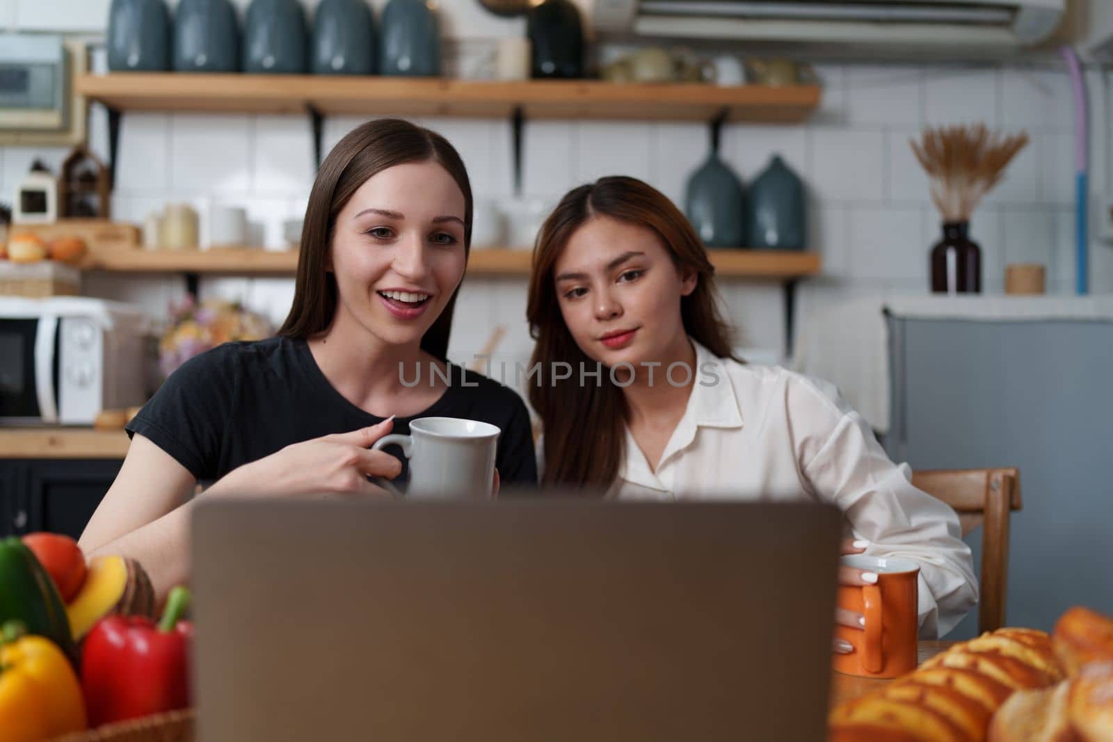 Beautiful woman and friend talking and using laptop at kitchen room. Girl friends couple living together at home