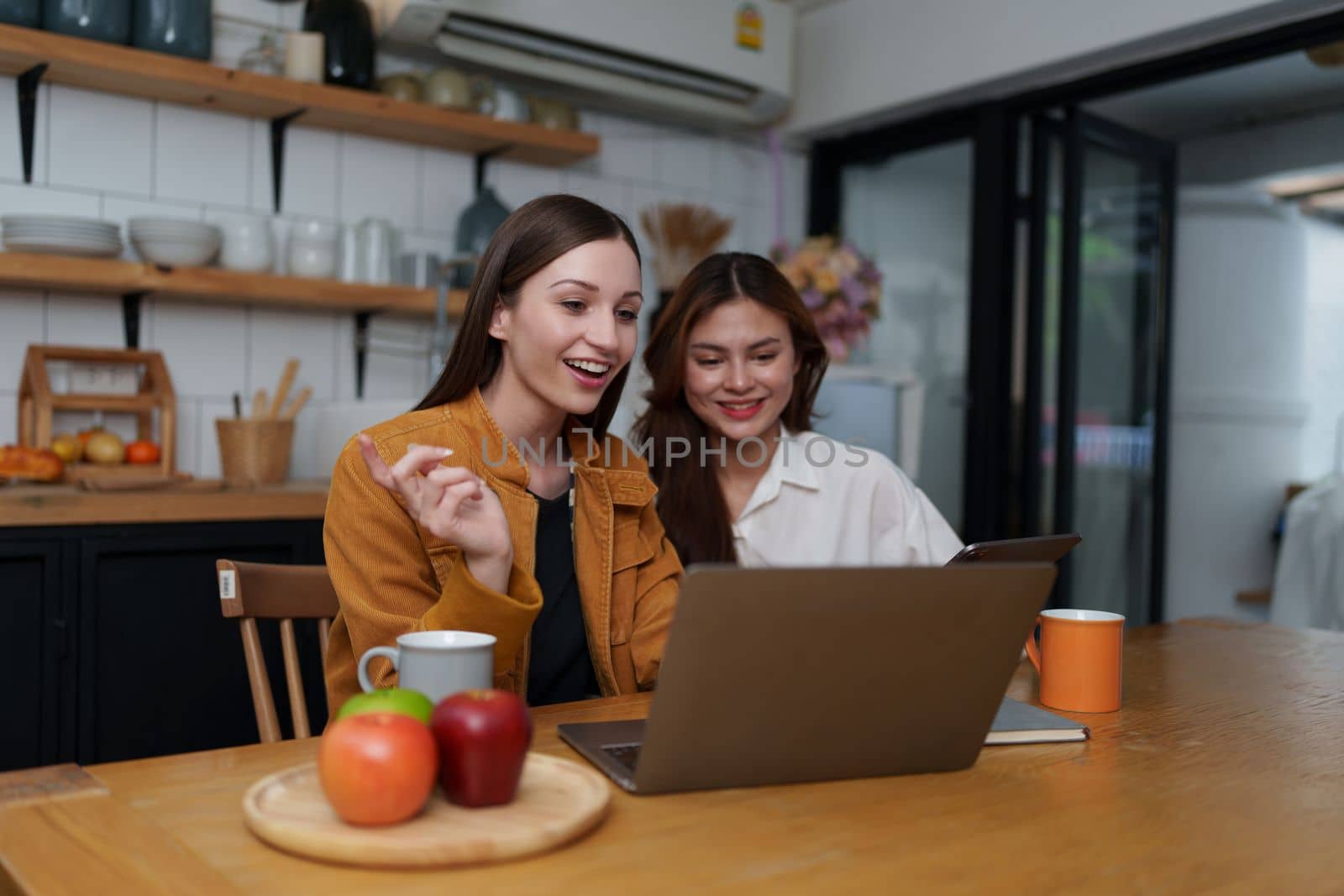 Beautiful woman and friend talking and using laptop at kitchen room. Girl friends couple living together at home