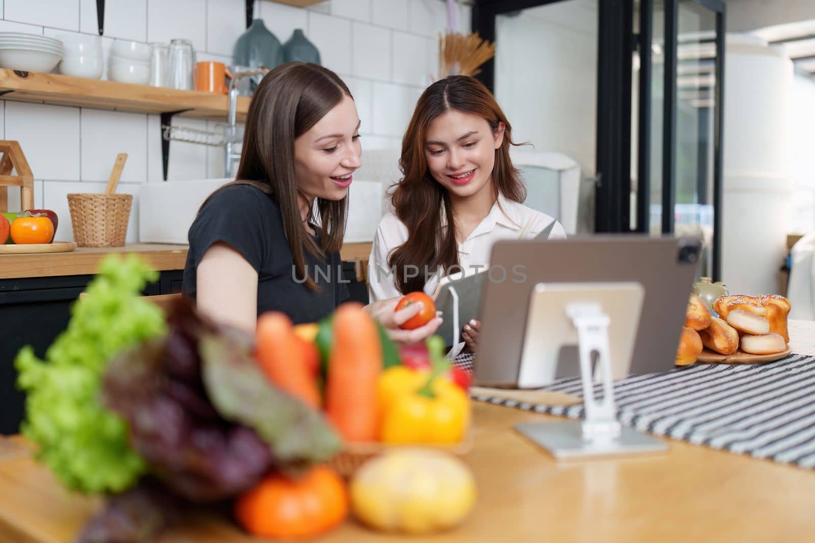 Beautiful woman and friend talking and using laptop at kitchen room. Girl friends couple living together at home
