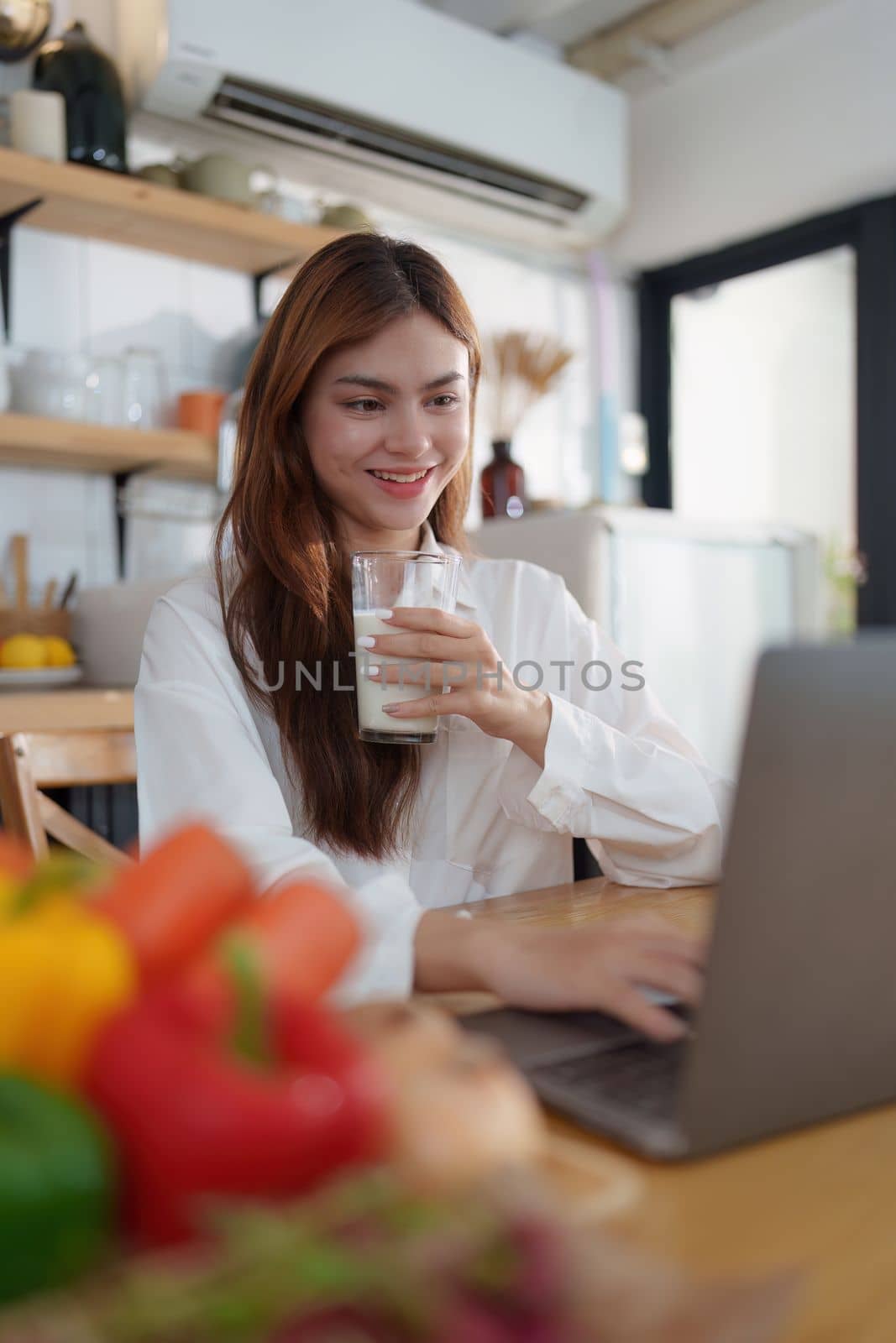 Portrait of a young cheerful asian woman drinking milk in kitchen room with healthy raw food. Vegetarianism, wellbeing and healthy lifestyle concept