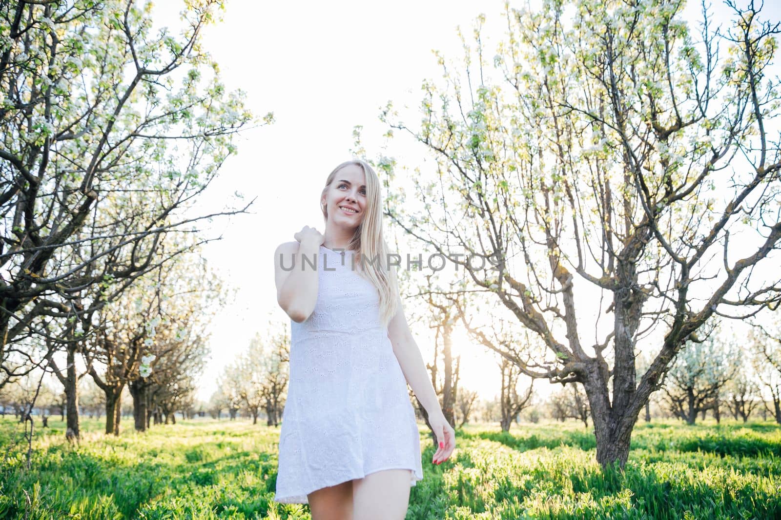 a blonde woman in white dress walks in park flowering trees garden