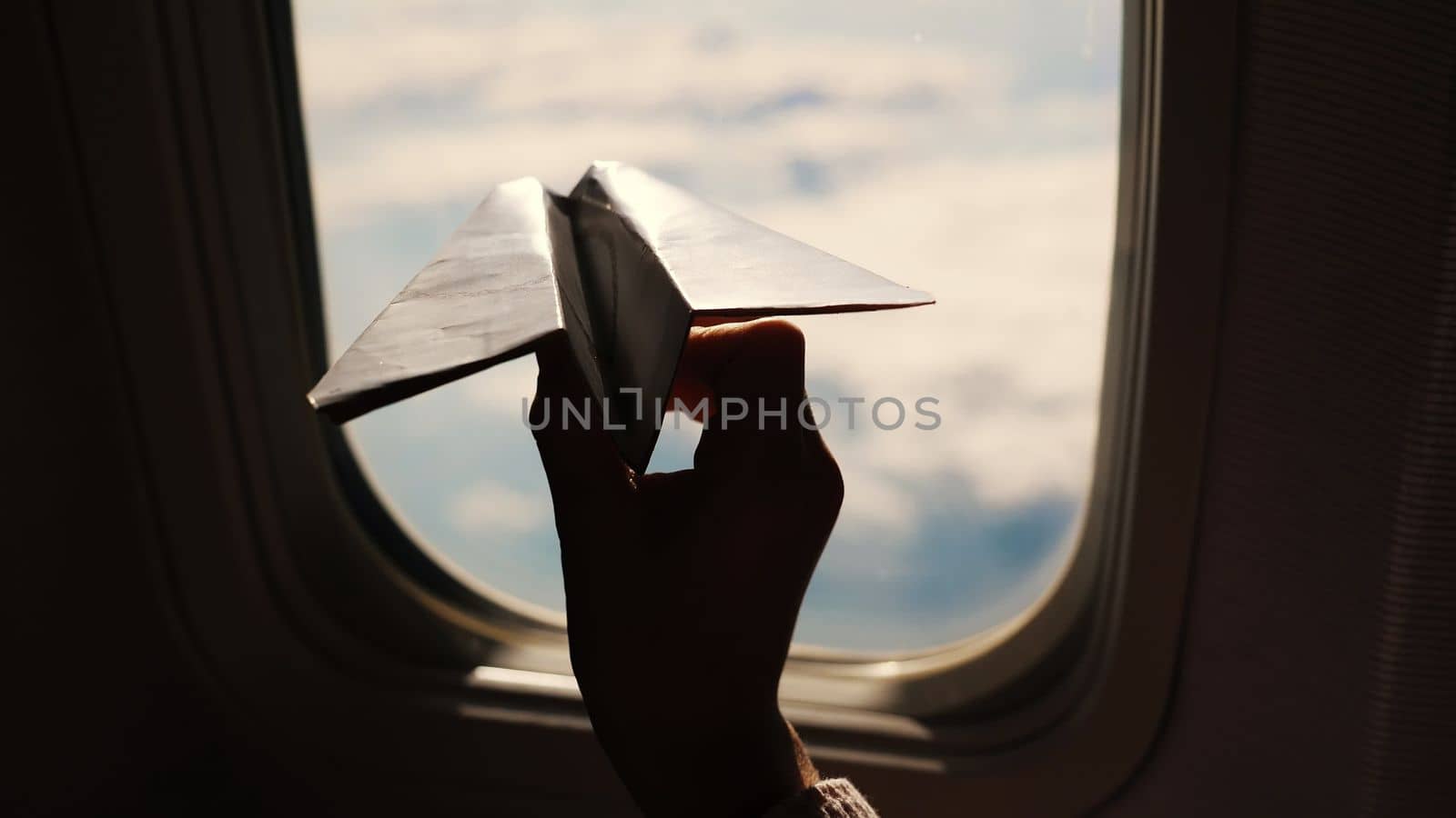 close-up. Silhouette of a child's hand with small paper plane against the background of airplane window. Child sitting by aircraft window and playing with little paper plane. during flight on airplane. High quality photo