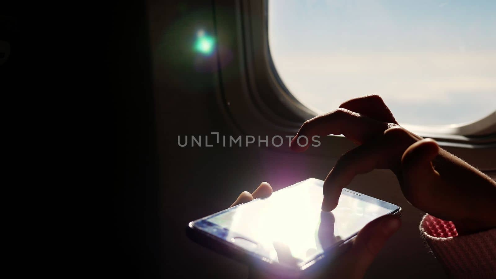 close-up. the sun's rays are reflected in the phone. dark silhouette of kid hands and mobile phone against airplane's illuminator. Child using, plays game on smartphone, tablet phone in airplane cabin. High quality photo