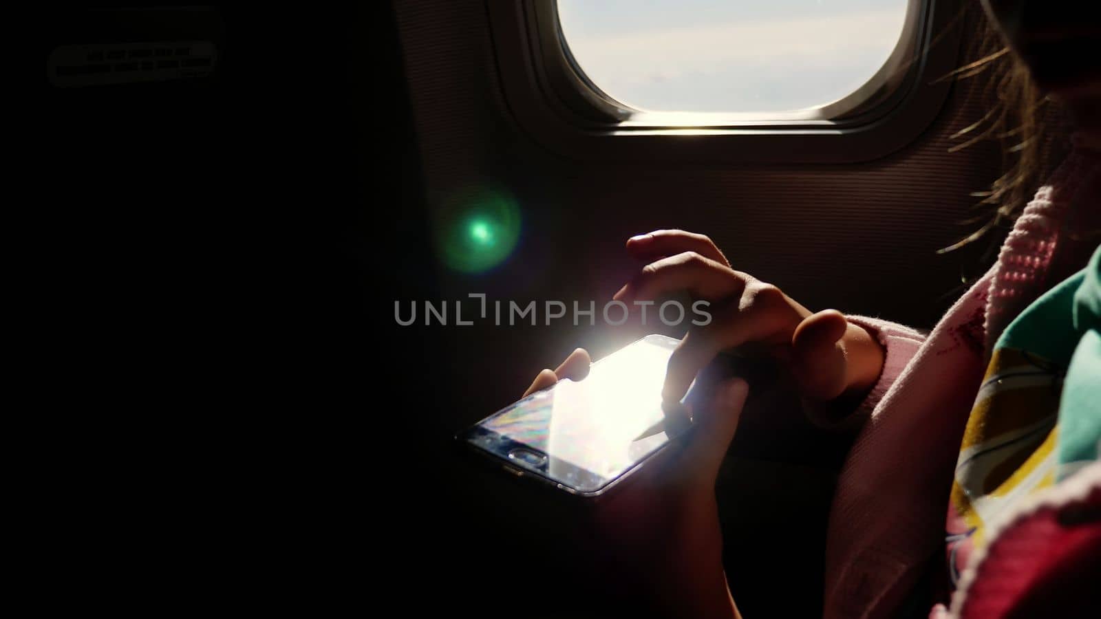 close-up. the sun's rays are reflected in the phone. dark silhouette of kid hands and mobile phone against airplane's illuminator. Child using, plays game on smartphone, tablet phone in airplane cabin. by djtreneryay