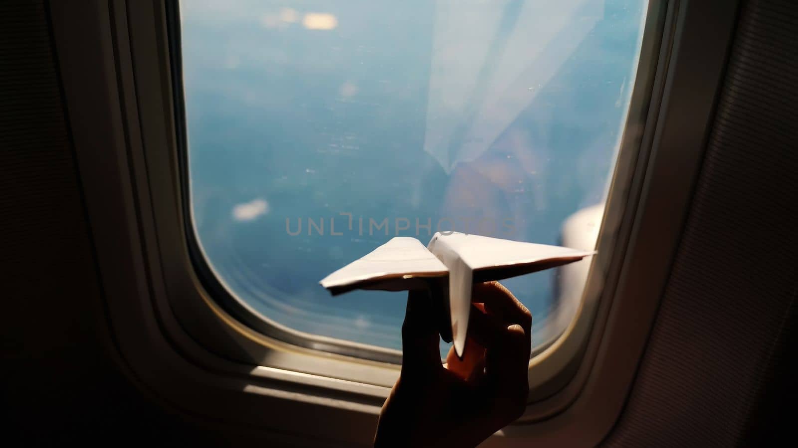 close-up. Silhouette of a child's hand with small paper plane against the background of airplane window. Child sitting by aircraft window and playing with little paper plane. during flight on airplane. High quality photo