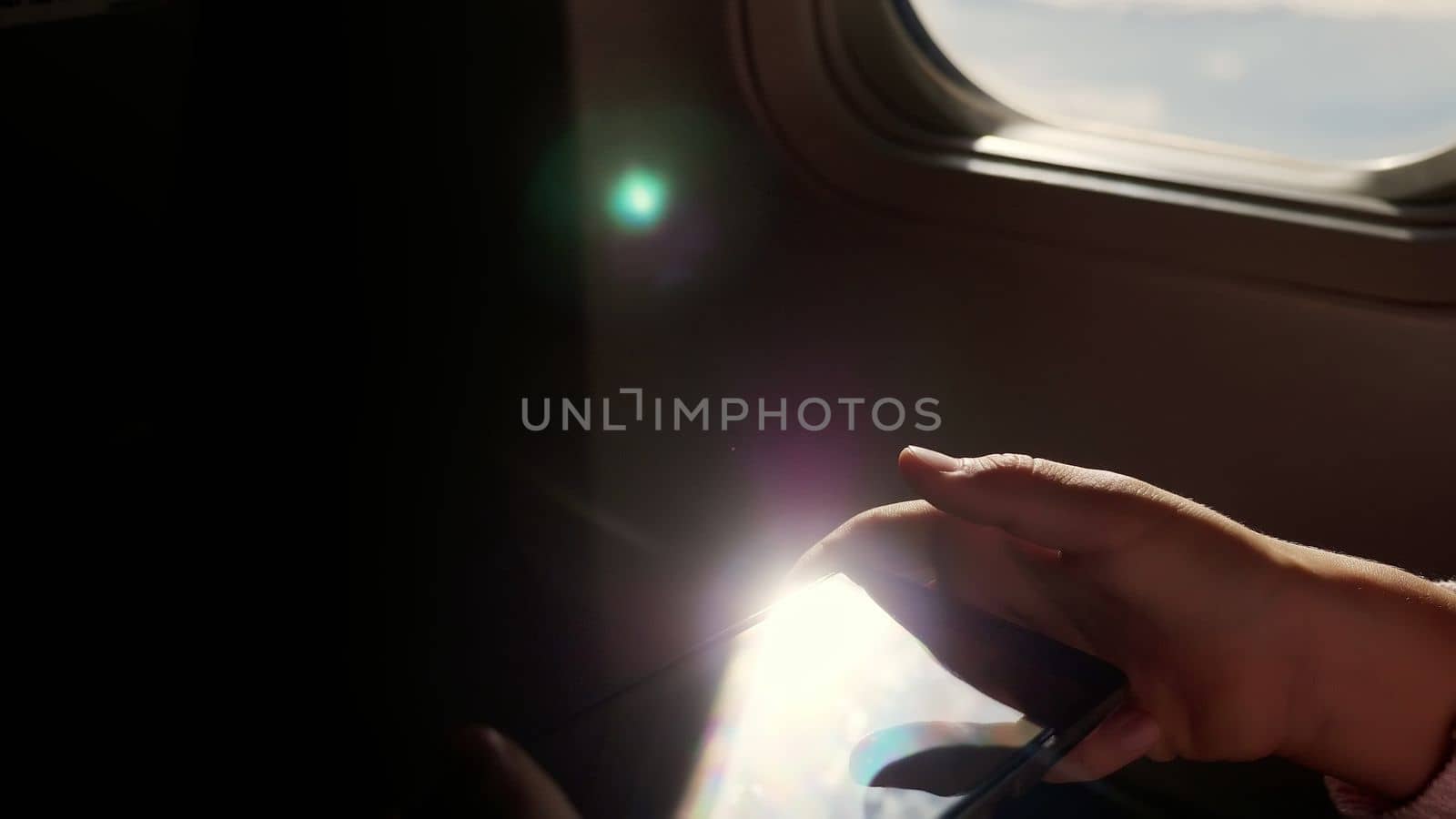 close-up. the sun's rays are reflected in the phone. dark silhouette of kid hands and mobile phone against airplane's illuminator. Child using, plays game on smartphone, tablet phone in airplane cabin. by djtreneryay