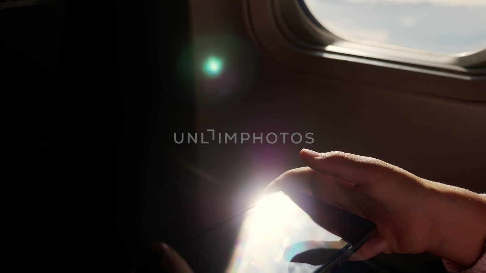 close-up. the sun's rays are reflected in the phone. dark silhouette of kid hands and mobile phone against airplane's illuminator. Child using, plays game on smartphone, tablet phone in airplane cabin. High quality photo