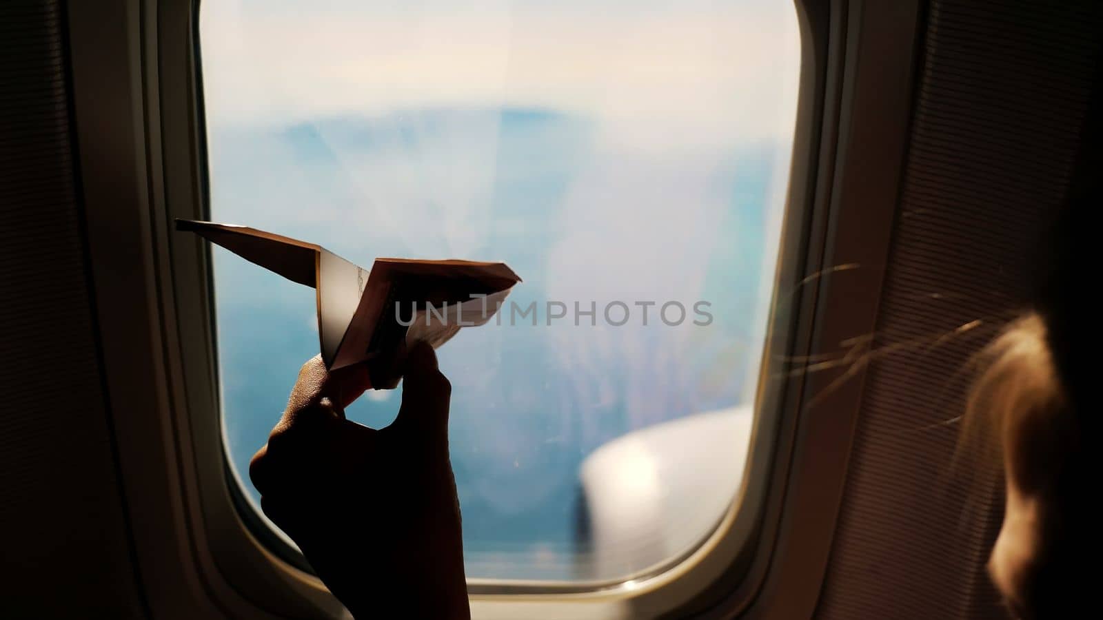 close-up. Silhouette of a child's hand with small paper plane against the background of airplane window. Child sitting by aircraft window and playing with little paper plane. during flight on airplane. High quality photo