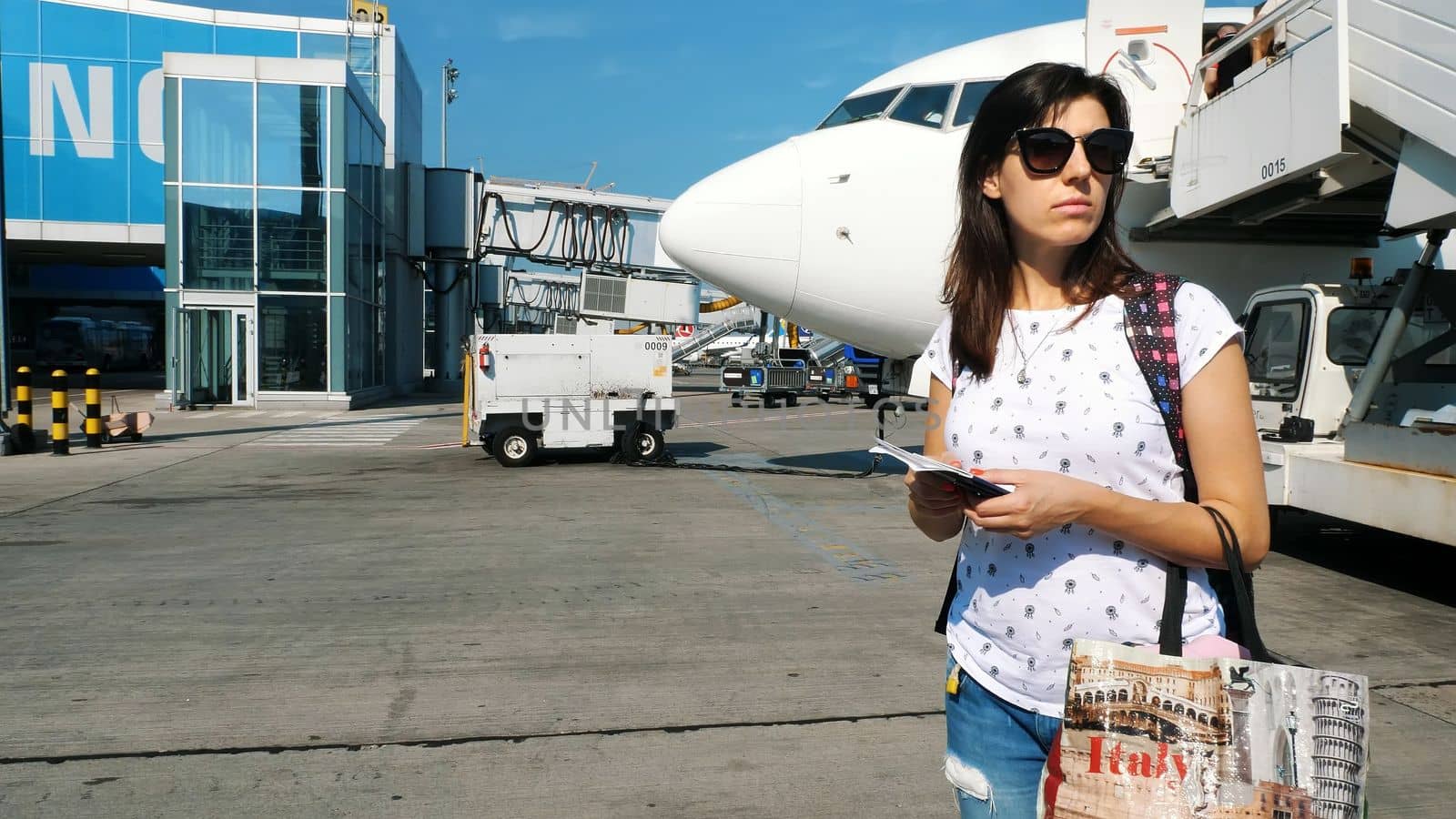 brunette woman in sun glasses, with backpack, holding Airline boarding pass tickets, and passport at airport, on background of the aircraft. summer. High quality photo