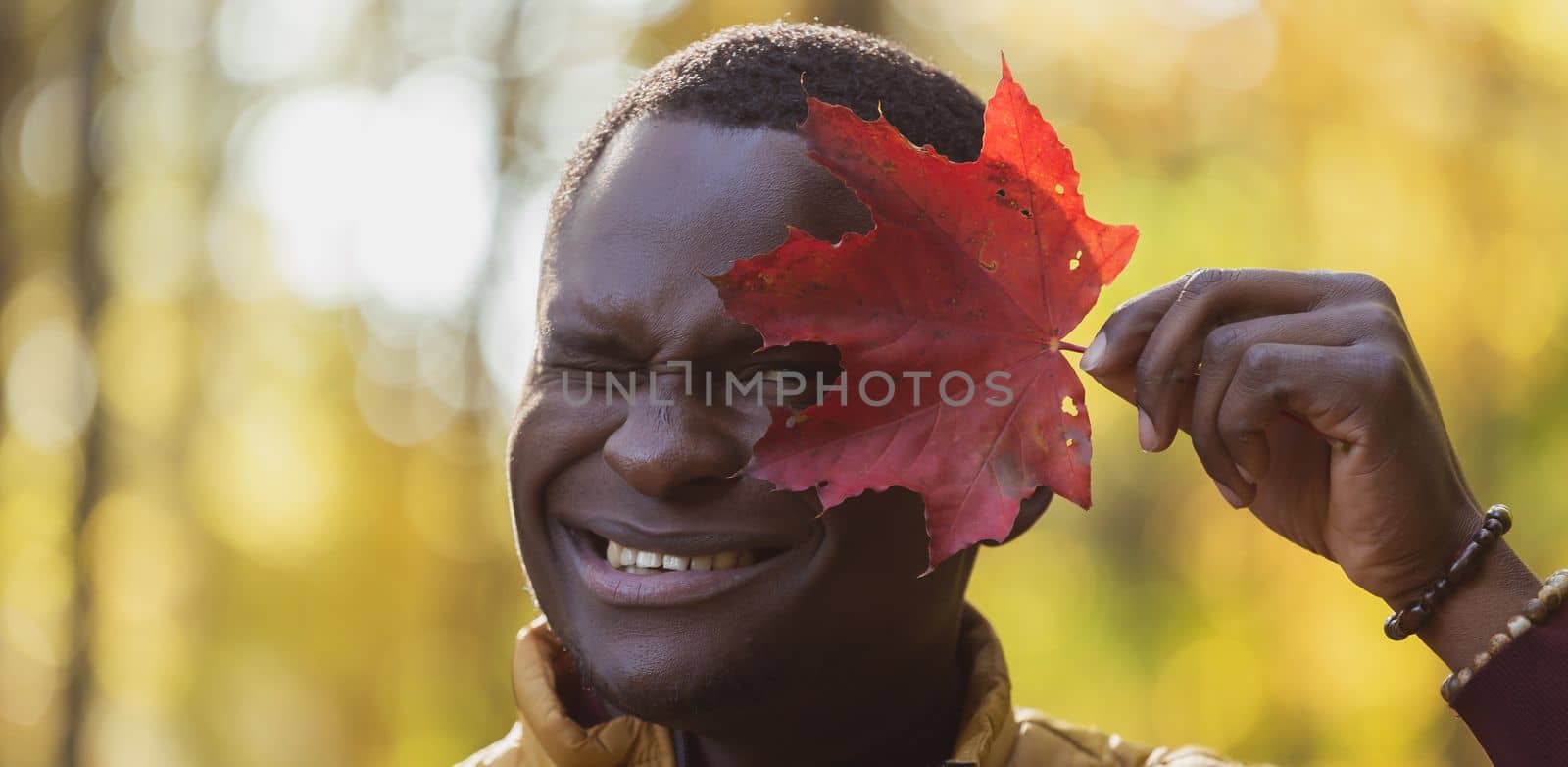 Banner Close up portrait of funny african american man covering his face with autumn maple leaf with copy space. Autumn nature. Seasonal fall fashion. by Satura86