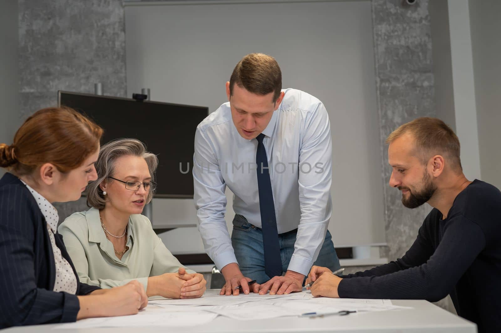 A caucasian man is standing making changes to a drawing, three colleagues are sitting at a table and listening to him