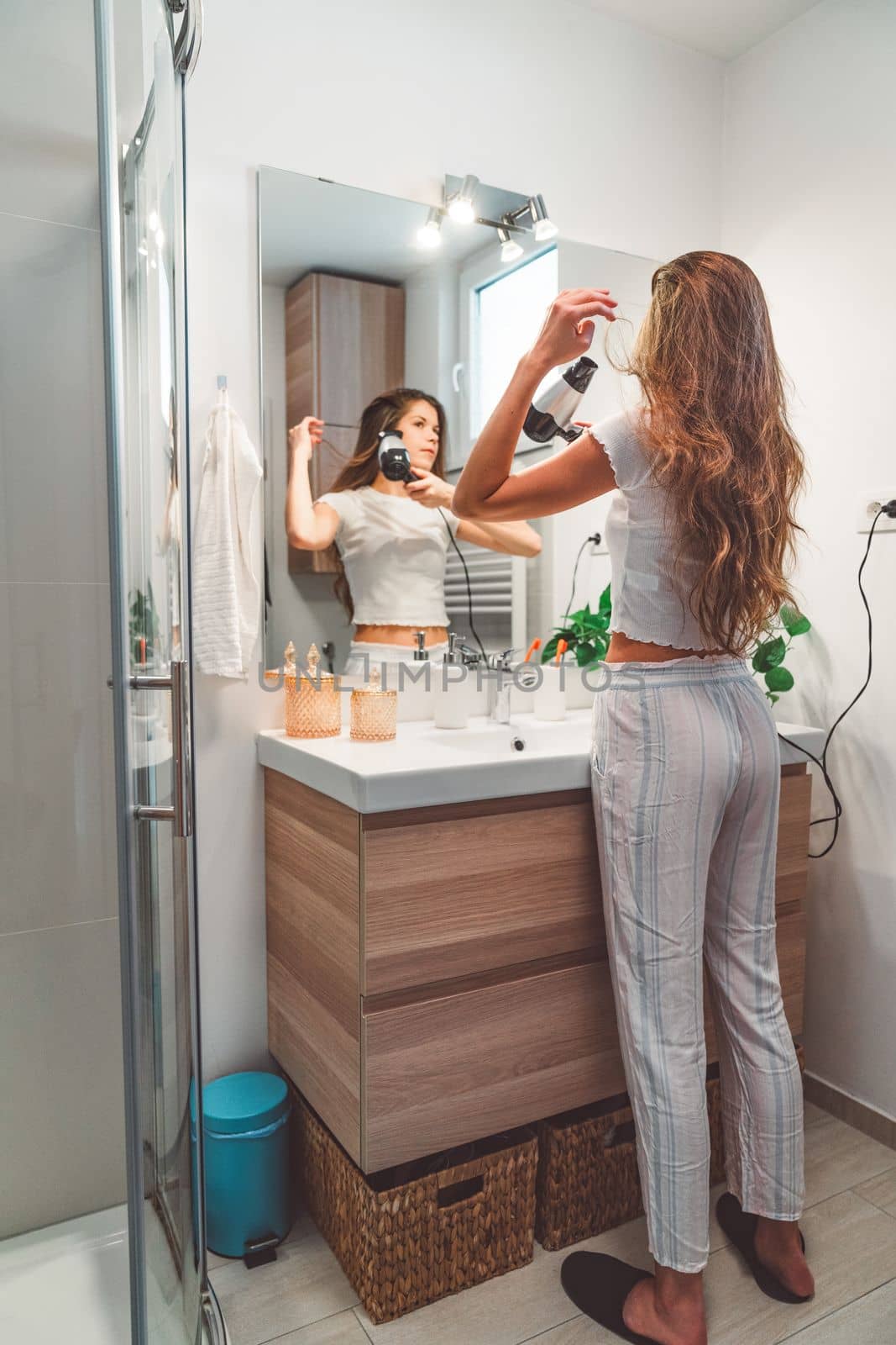 Vertical photo of young caucasian woman standing at the bathroom sink drying her hair with a hair dryer by VisualProductions