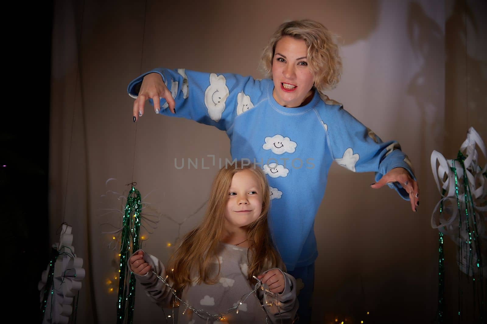 Cute mother and daughter in pajamas having fun in the room with Christmas garlands and white background. The tradition of decorating the house for the holidays. Happy childhood and motherhood by keleny