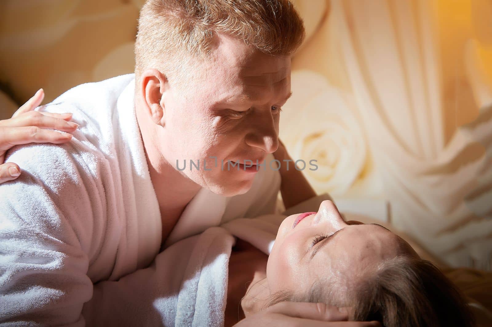 An adult couple of newlyweds in in white robes in a hotel room after the wedding. A guy and a girl, a man and a woman in beautiful room. The concept of hugs, love and care