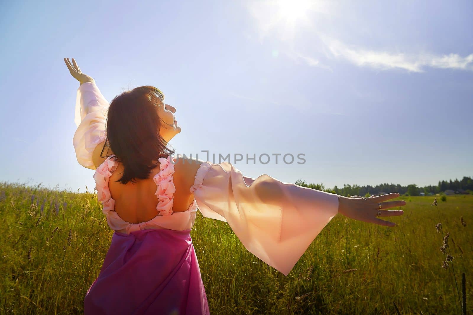 Beautiful girl in lush pink ball gown in green field during blooming of flowers and blue sky on background. Model posing on nature landscape as princess from fary tale