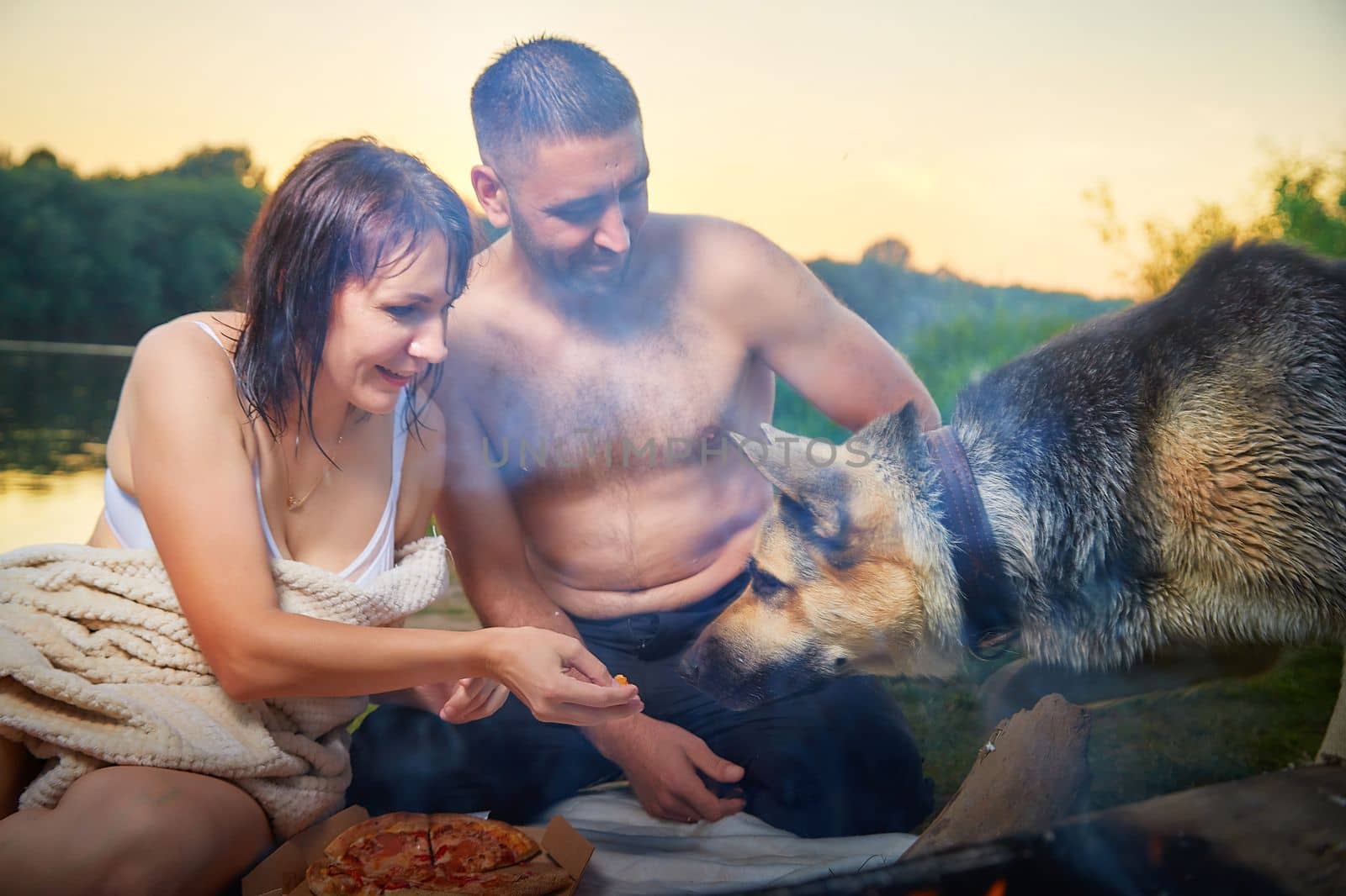 Happy wet couple relaxin, having fun and hugs with big dog near fire in camping on nature in summer sunny evening in sunset. Family or lovers have date and rest outdoor. Concept of love. Partial focus