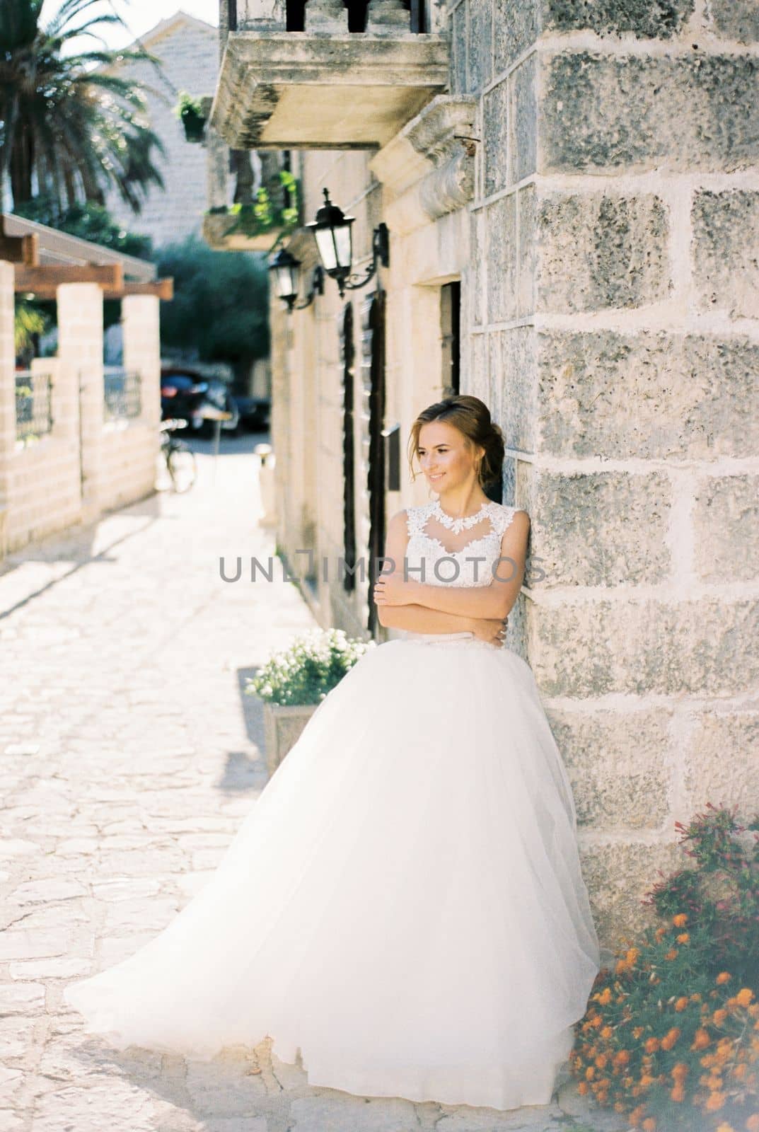 Bride with her arms crossed on her chest stands leaning against a stone house. High quality photo
