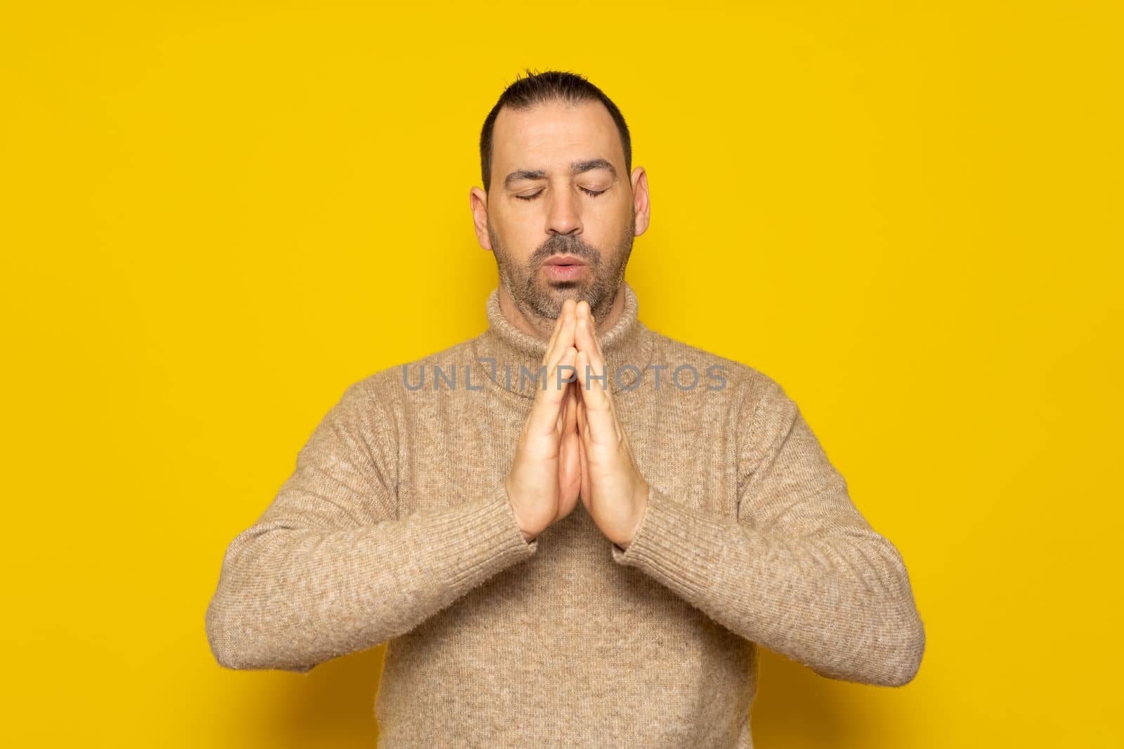 Bearded hispanic man wearing beige turtleneck isolated over yellow background, holding hands in prayer or meditation, looking relaxed and calm, dreaming and hoping for the best