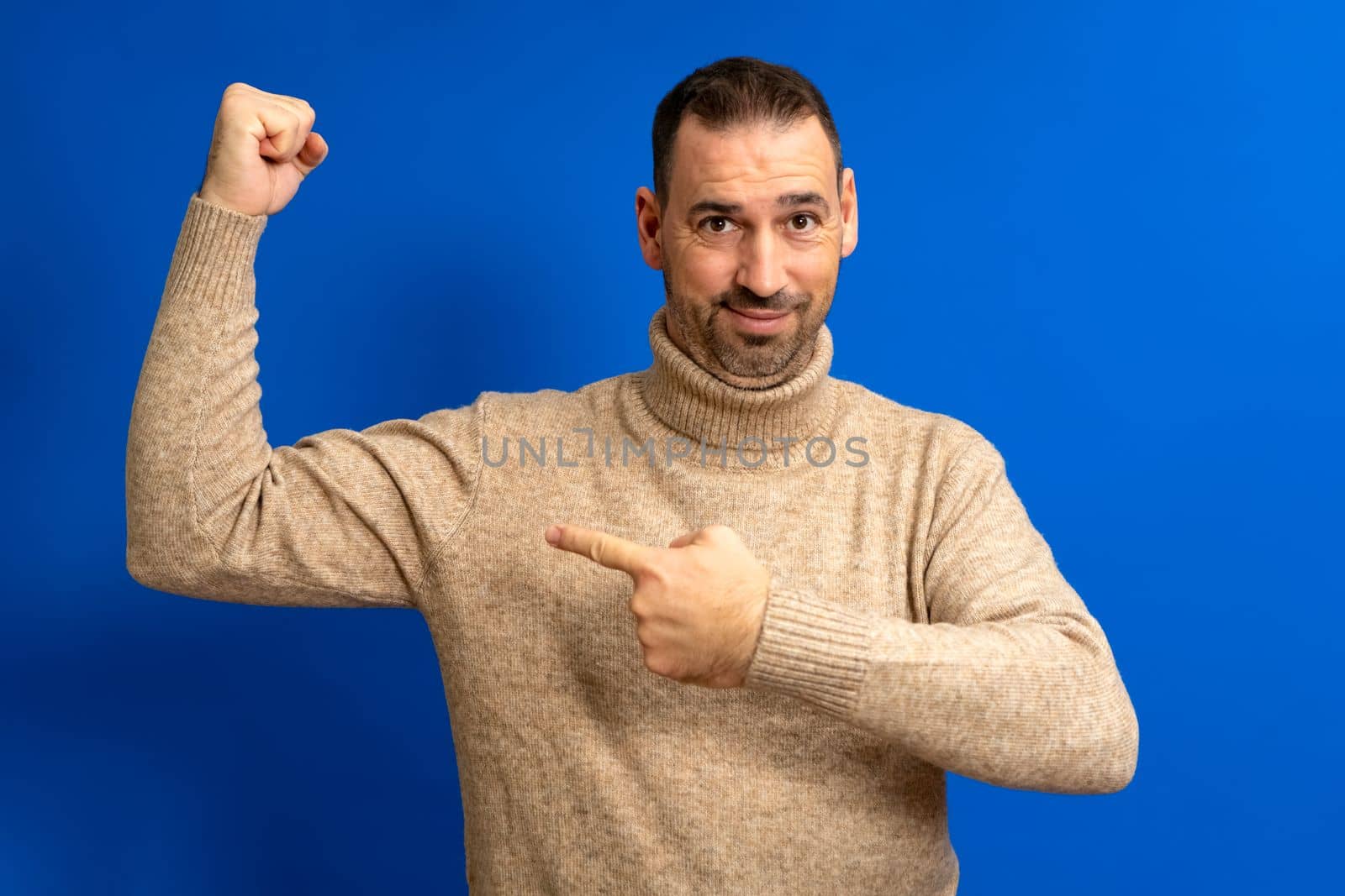 Portrait latin bearded man pointing finger on his arm muscle, discount, looking at camera with satisfied face. Indoor studio shot isolated on blue background.