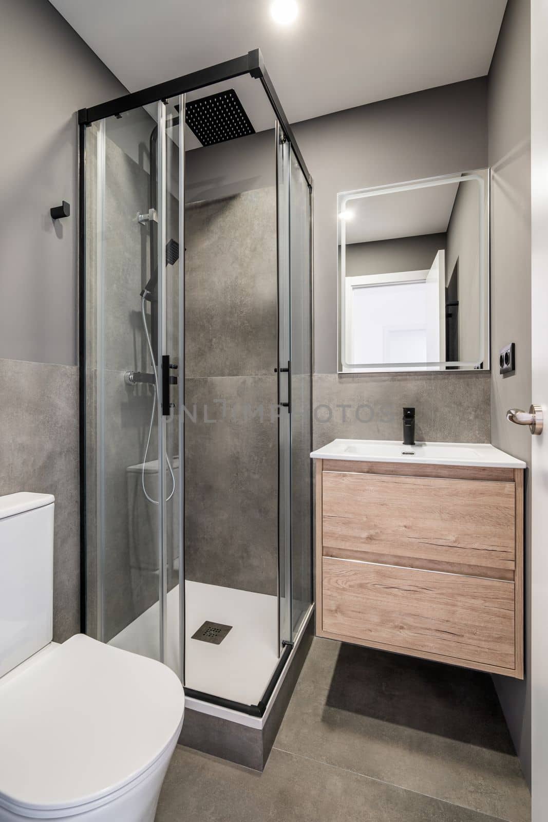 Small bathroom with bright lighting from white ceiling. In corner is shower with glass sliding doors. Toilet bowl and sink in white ceramic. On wall is designer mirror with reflection of front door