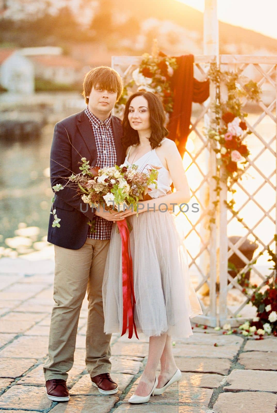 Groom and bride with a bouquet of flowers stand near the wedding screen on the pier. High quality photo