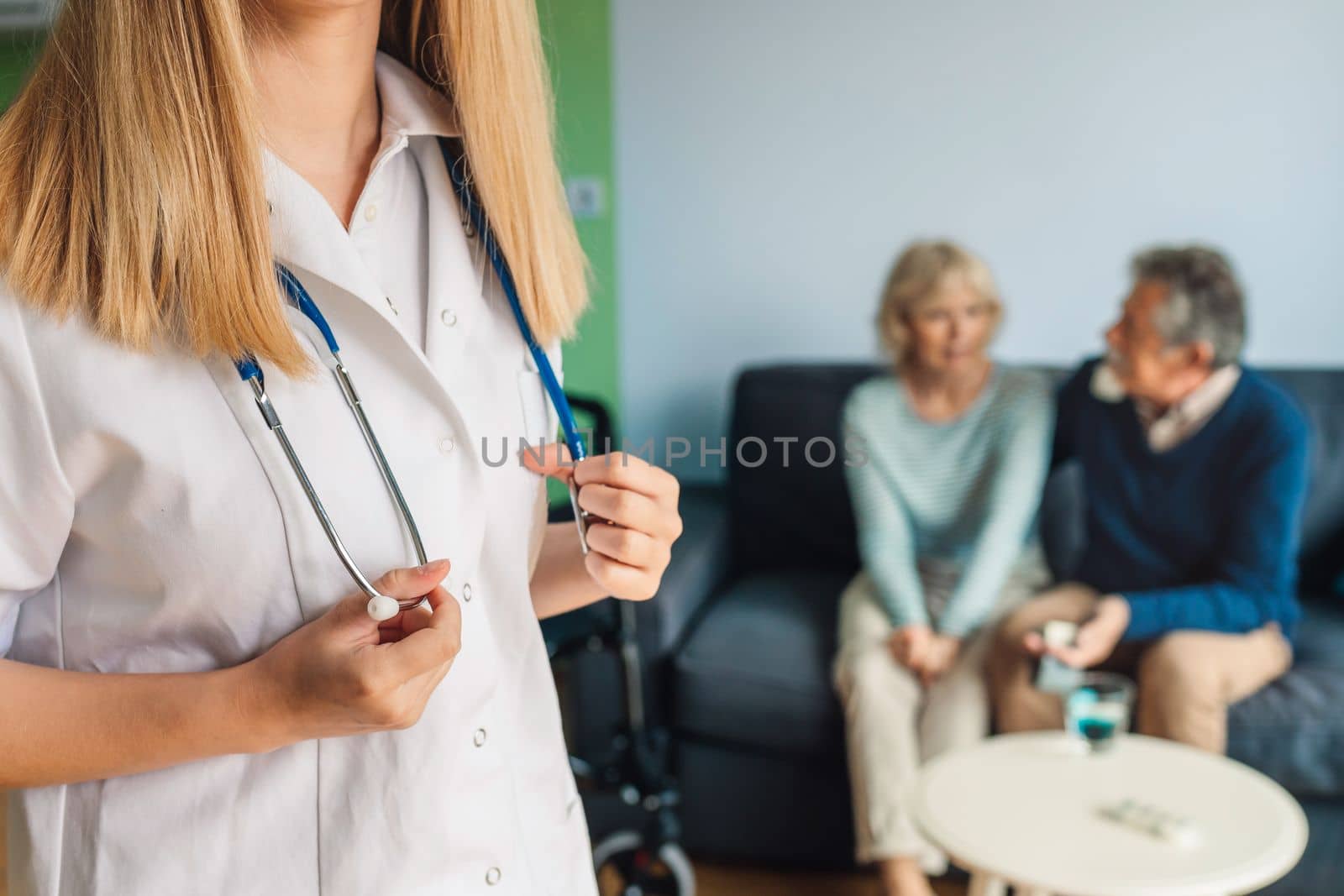 Unrecognizable woman nurse holding for her stethoscope while senior couple is sitting on the couch blurred in the background by VisualProductions