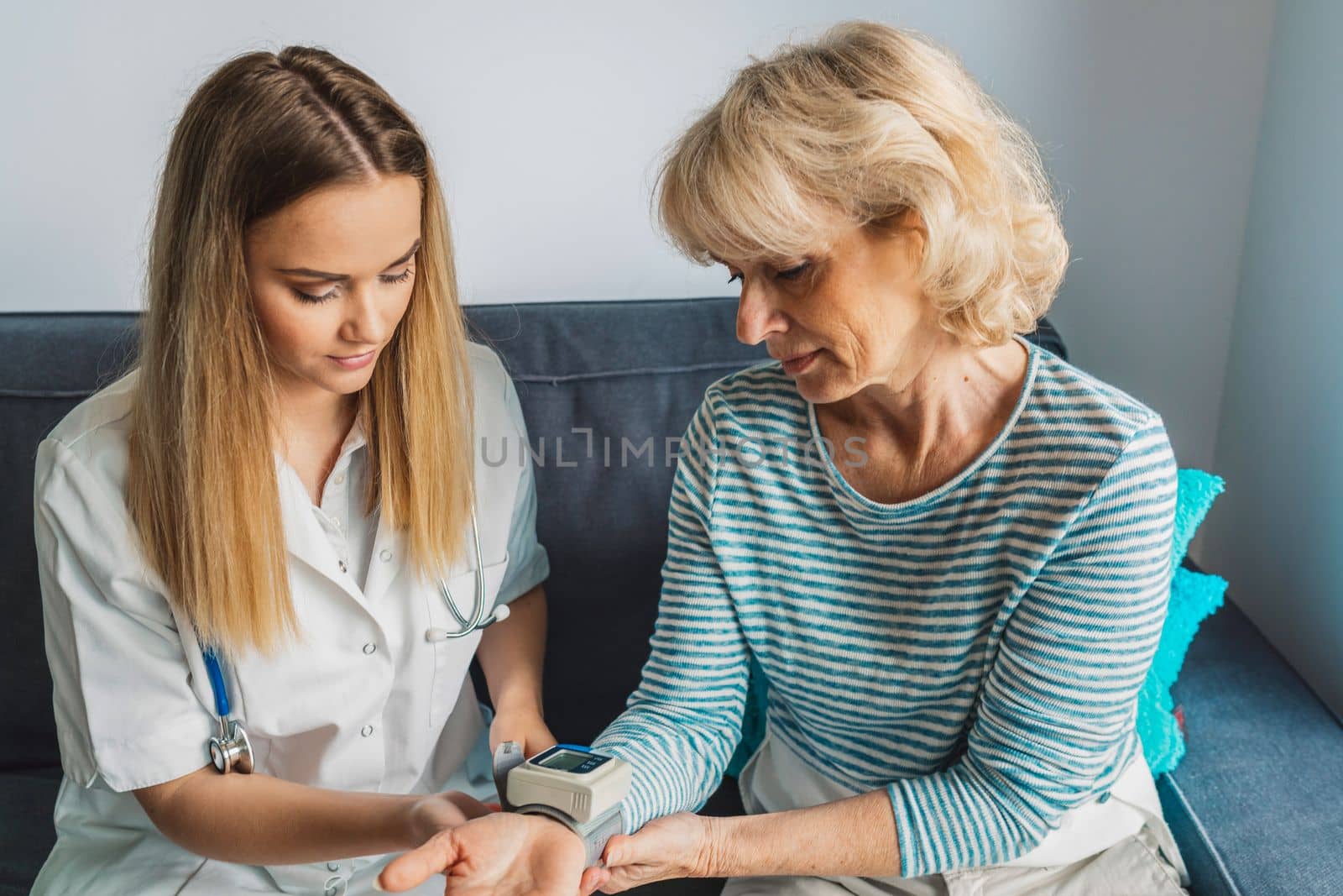 Nurse sitting down on the couch with elderly lady, while taking her blood pressure by VisualProductions