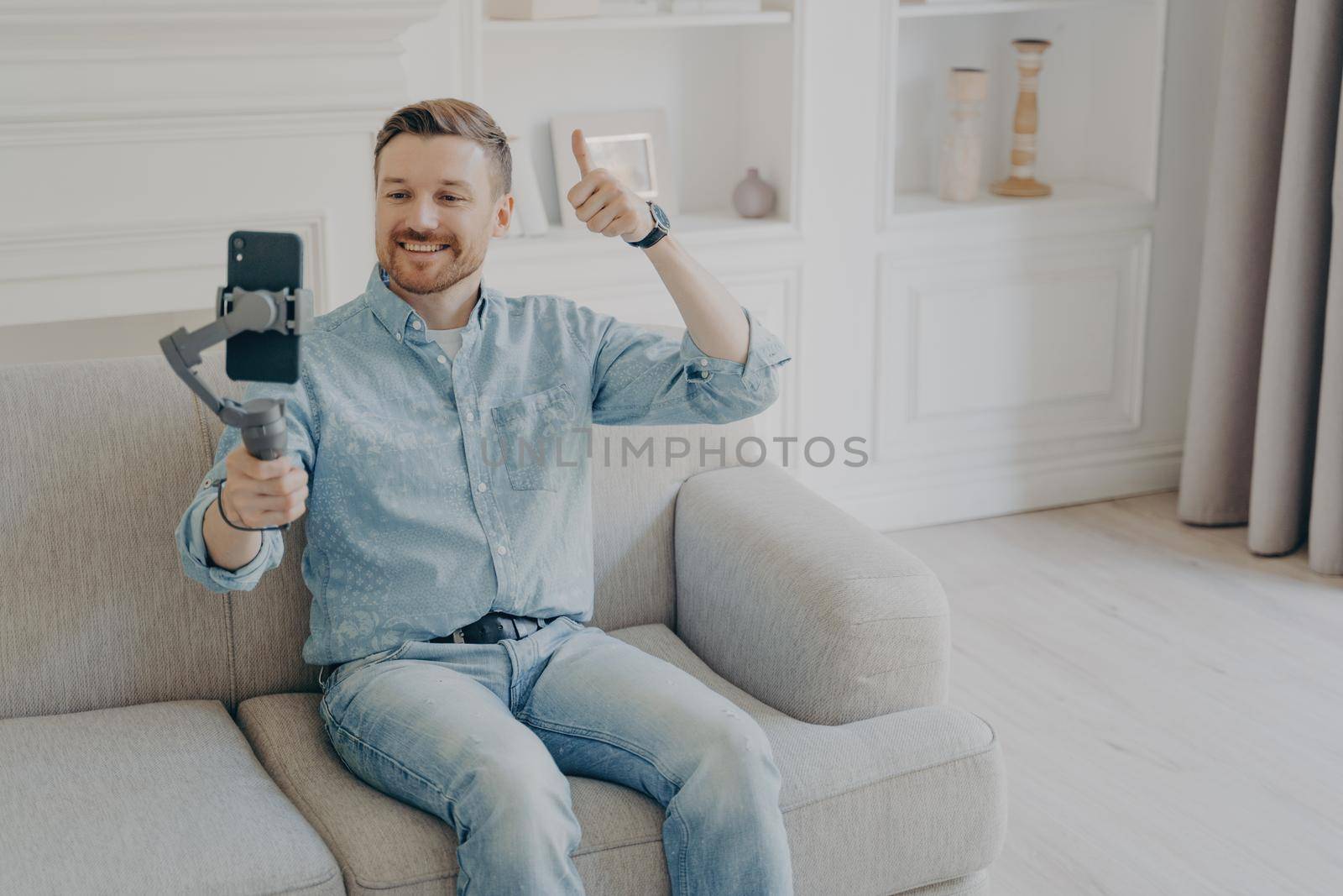 Positive smiling young man dressed in casual clothes showing thumbs up gesture in video chat, holding phone using gimbal, sitting alone on beige couch in light themed living room