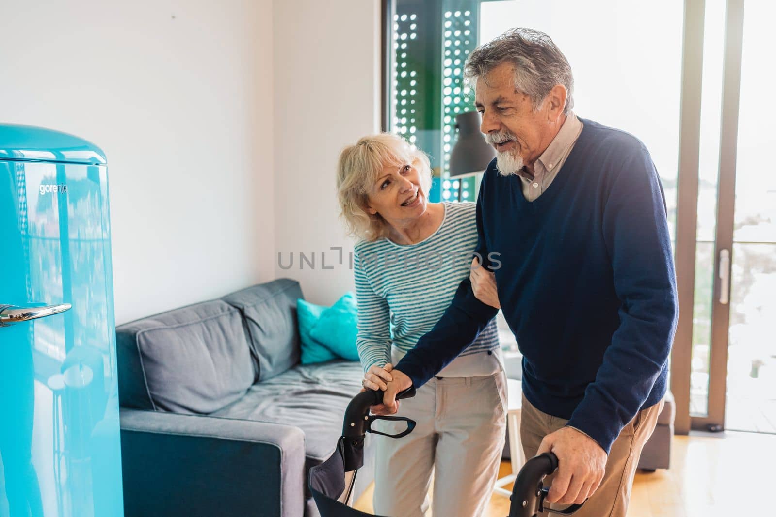 Senior woman helping senior man walking inside the apartment at home. High quality photo