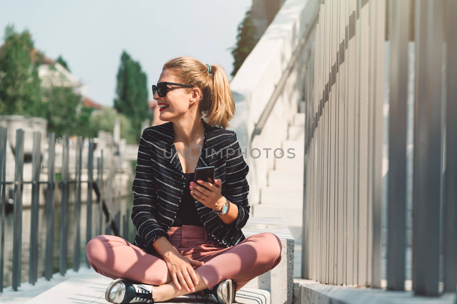 Daily life in the city, woman sitting on the sun outside, taking on speaker on the phone by VisualProductions