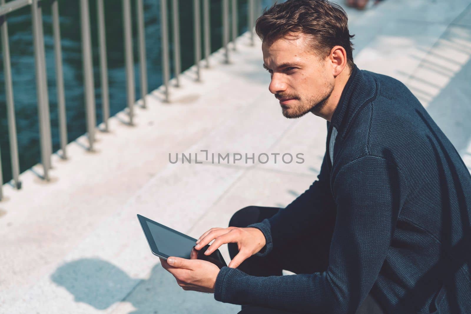 Caucasian man sitting by the river in the city working on his tablet during lunch time by VisualProductions