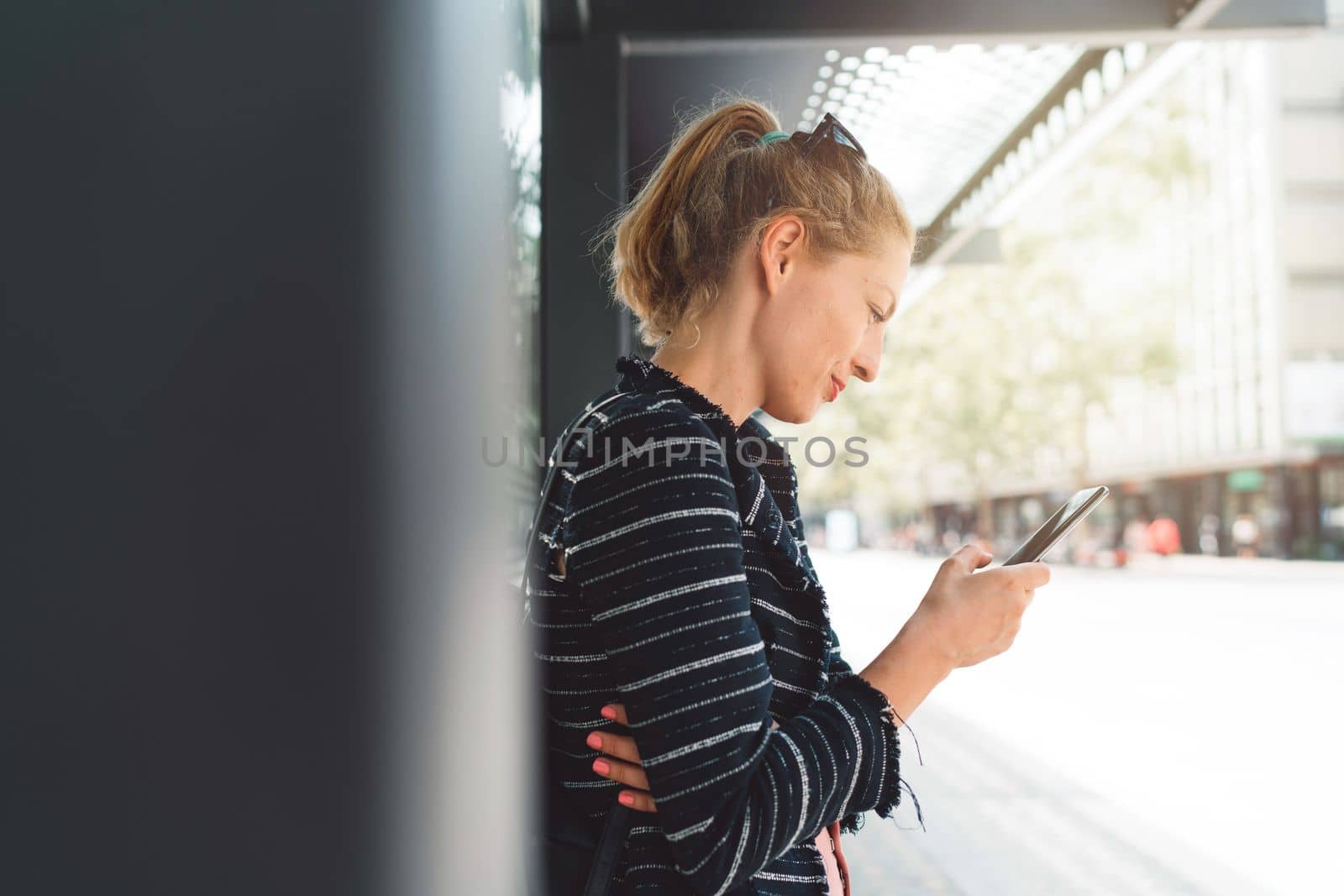 Side profile of caucasian blonde woman staring at her phone while waiting for her bus by VisualProductions