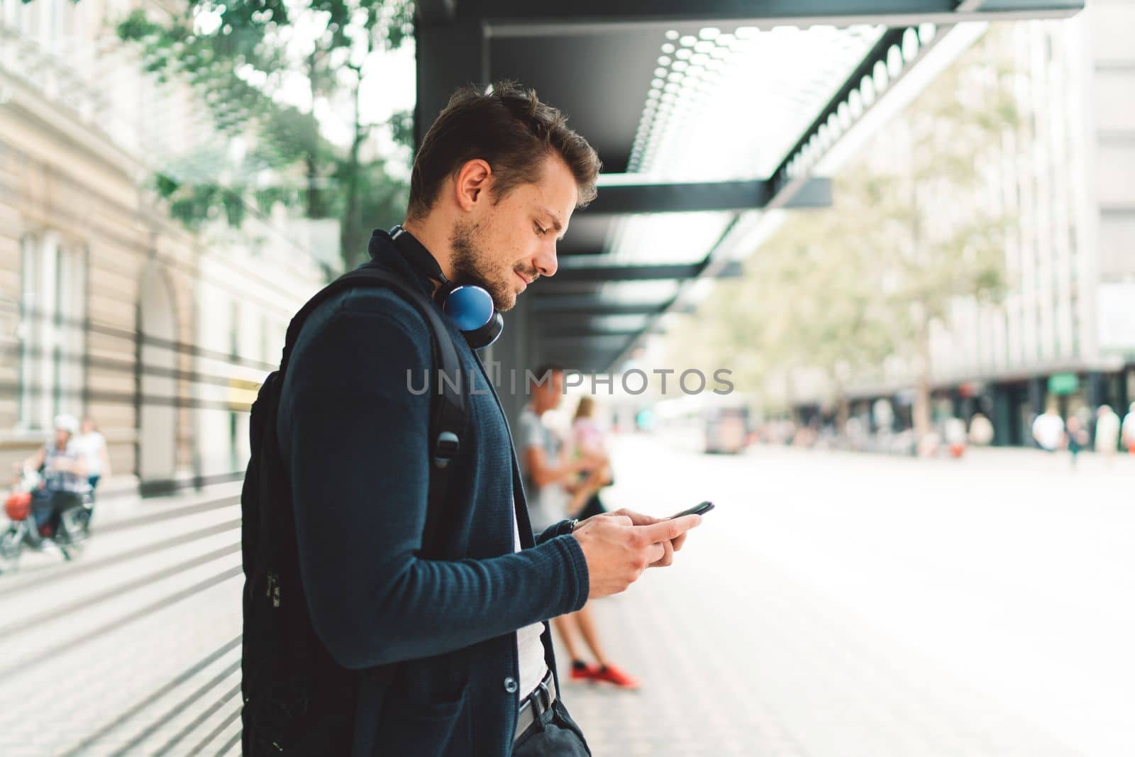 Side view of young man looking down at his phone while waiting for his bus to come by VisualProductions