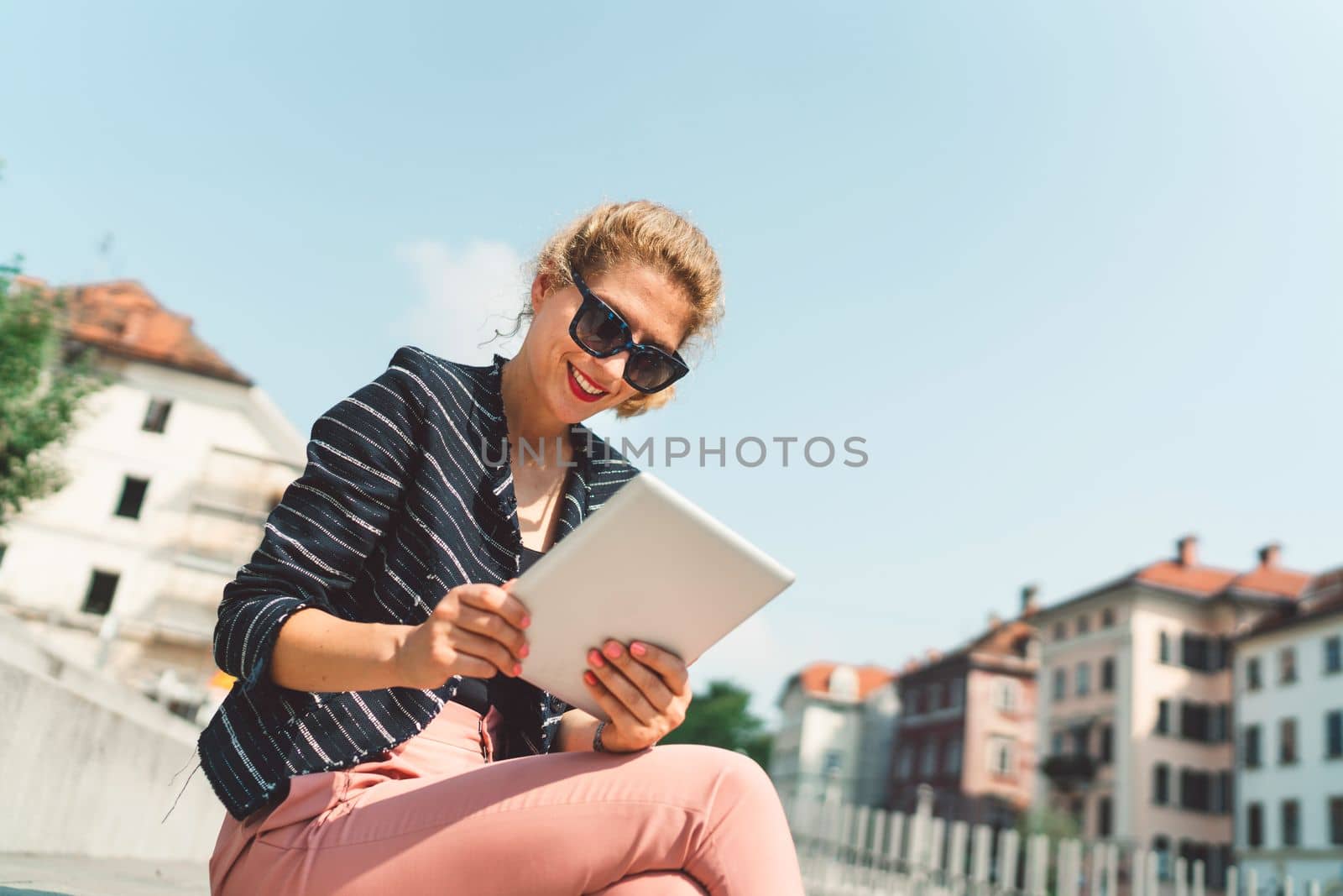 Low angle view smiling caucasian woman sitting on the sun working on her digital tablet by VisualProductions