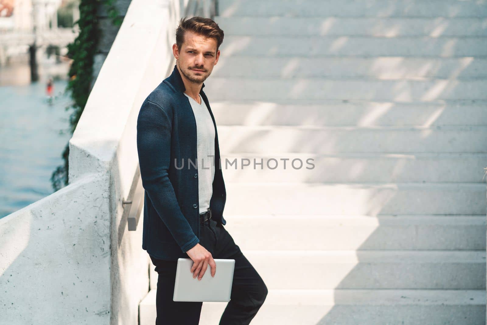 Side view of caucasian man standing on the stairs holding a tablet in one hand, sunny day outside by VisualProductions