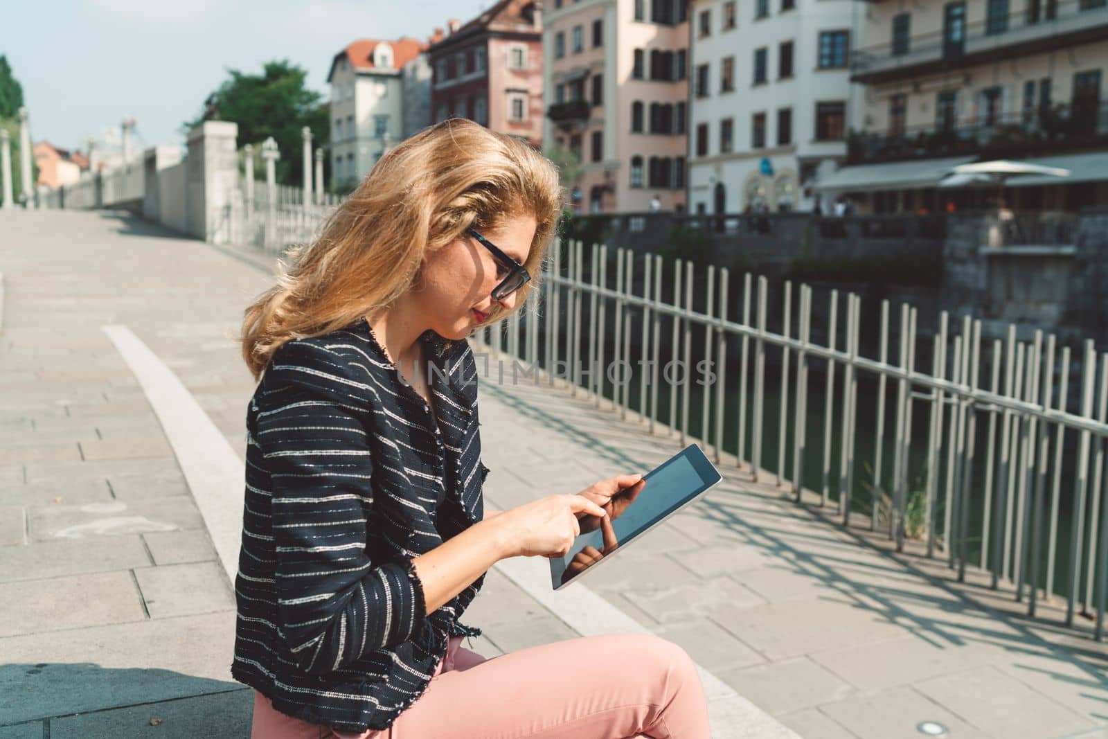 Beautiful woman with blonde hair looking down at the tablet while sitting on the sun outside int he city by VisualProductions