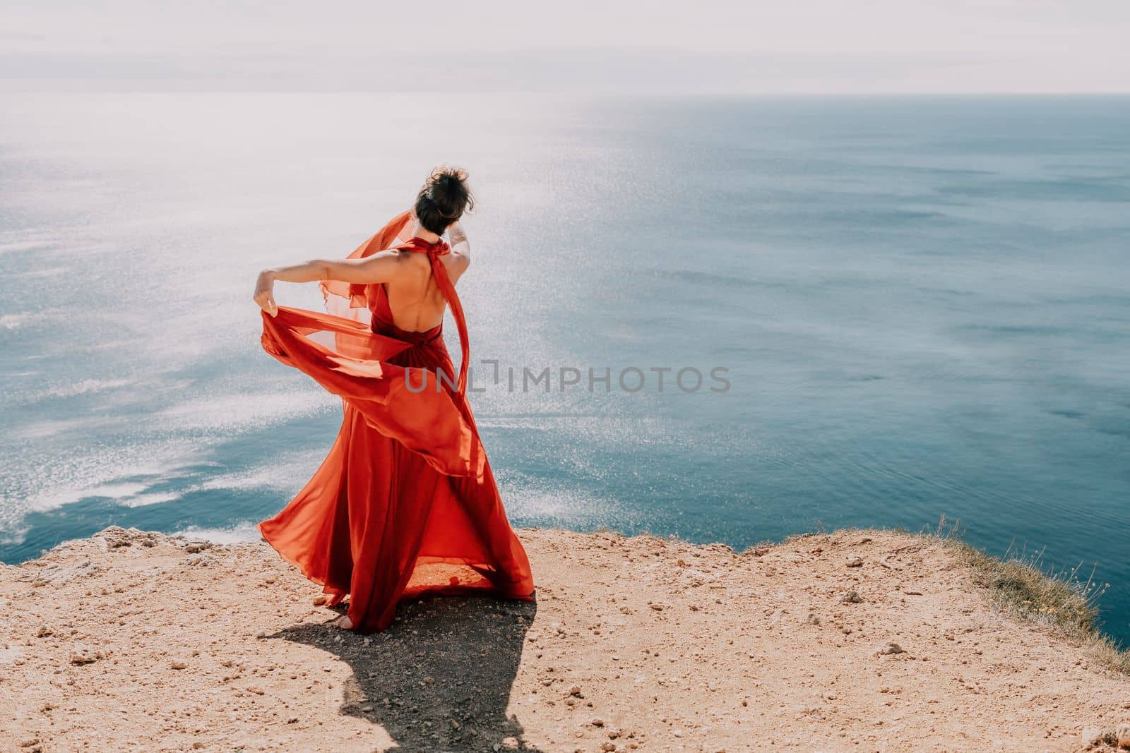 Side view a Young beautiful sensual woman in a red long dress posing on a rock high above the sea during sunrise. Girl on the nature on blue sky background. Fashion photo.