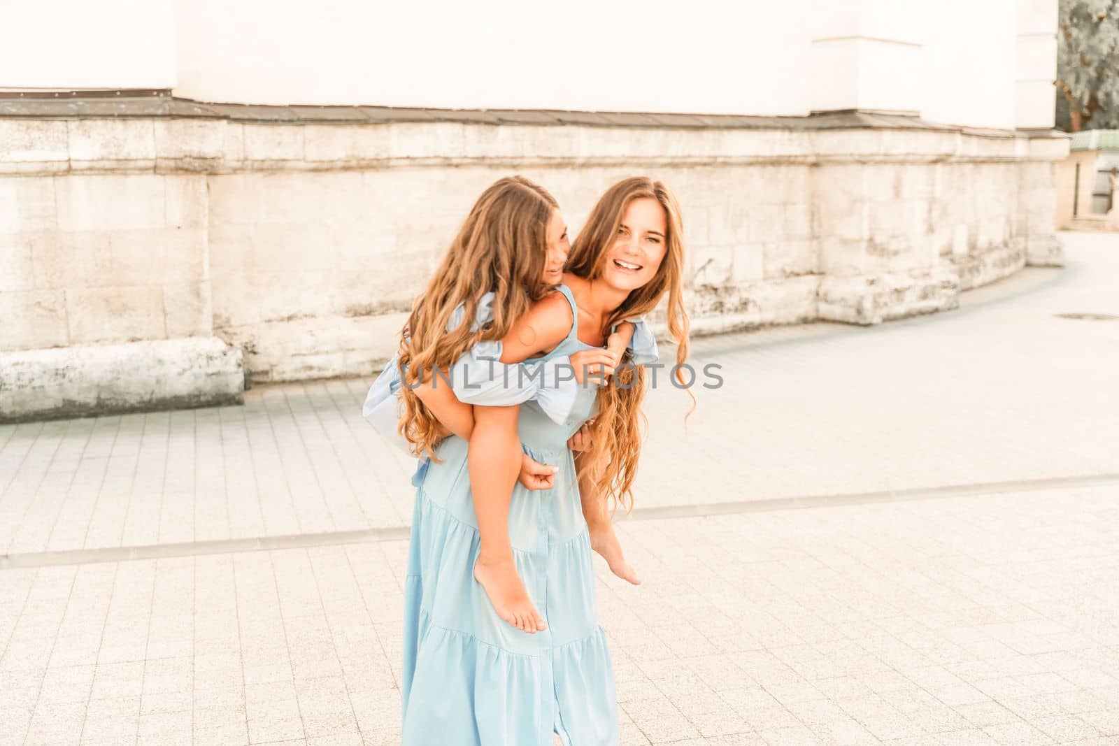 Mother of the daughter walks playing. Mother holds the girl on her back, holding her legs, and her daughter hugs her by the shoulders. Dressed in blue dresses