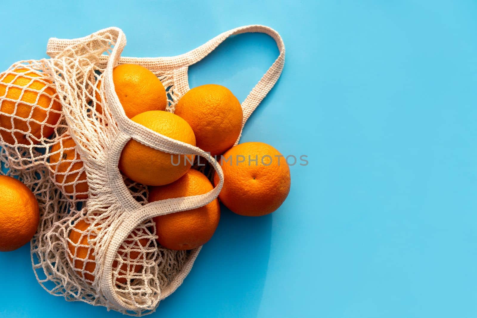 Mesh white cotton reusable bag with oranges on a blue background. Environmentally friendly shopping, the environment and the concept of healthy foods, citrus fruits with vitamin C. Place for text.