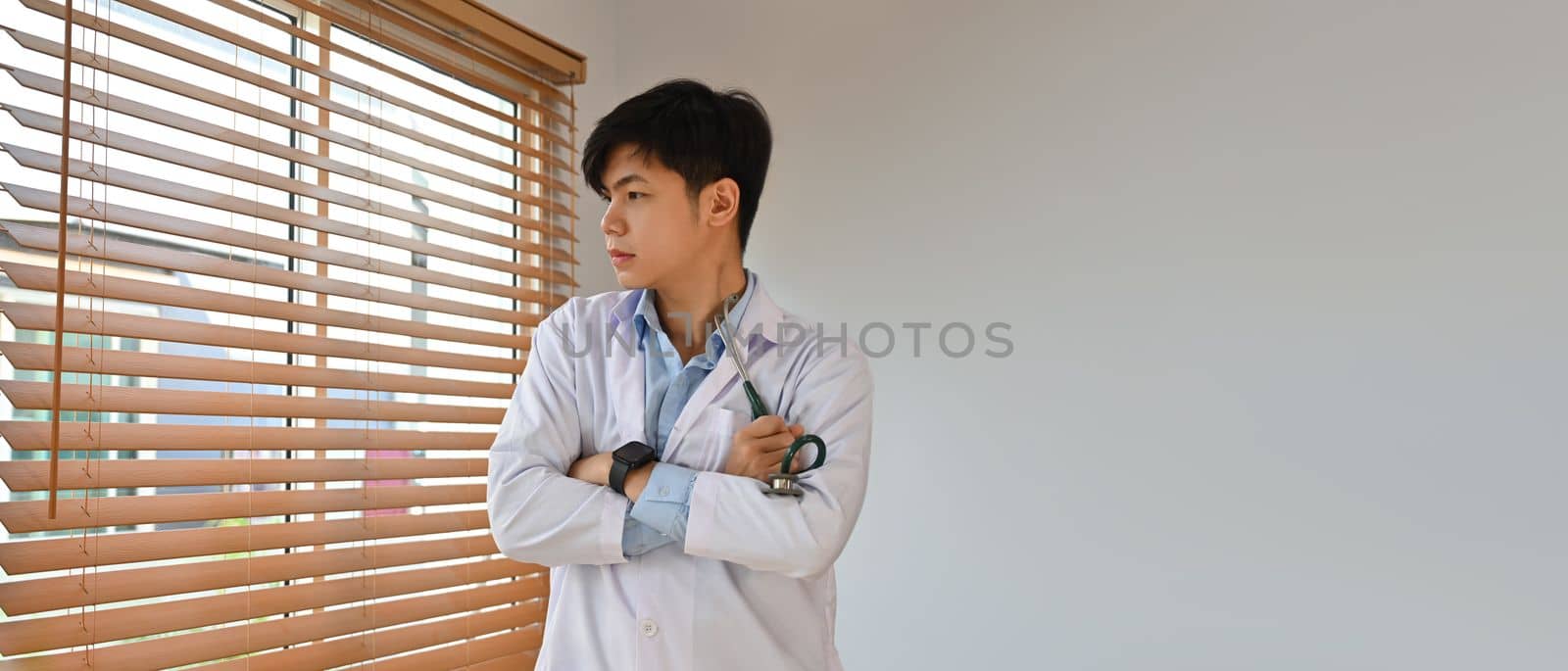 Banner panorama of thoughtful doctor wearing white uniform standing with arms crossed and looking through office window.