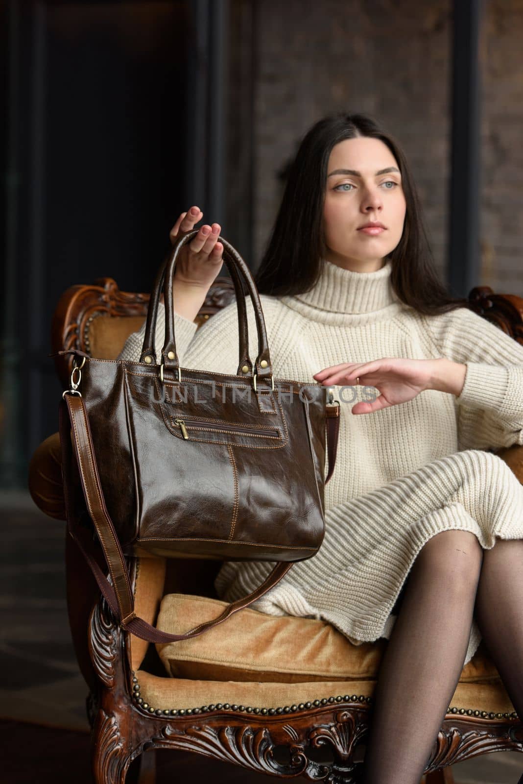 a brunette girl in a knitted beige dress poses while sitting with a shiny leather bag in her hands